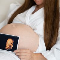 Unrecognizable pregnant woman wearing robe at home caressing,stroking belly, baby bump, holding ultrasound sonogram photo.Cropped, close-up shot.Happy,natural,calm, healthy pregnancy.